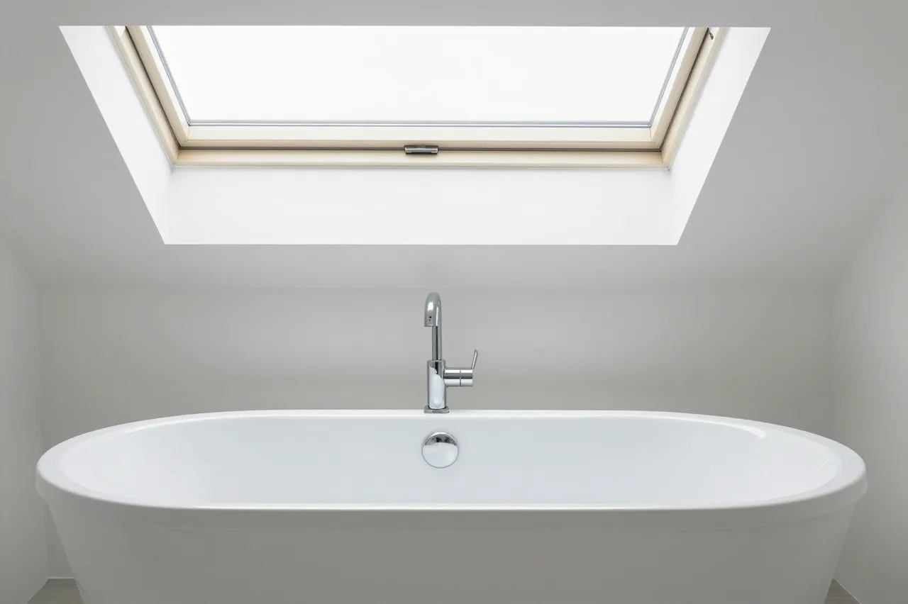 White freestanding soaking bathtub sits directly beneath a ceiling skylight in a minimalist white bathroom