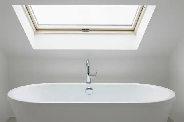 White freestanding soaking bathtub sits directly beneath a ceiling skylight in a minimalist white bathroom