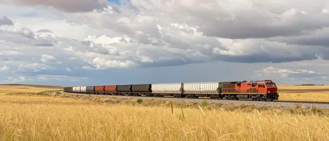 A long freight train curves through dry golden grassland under a cloudy wide sky