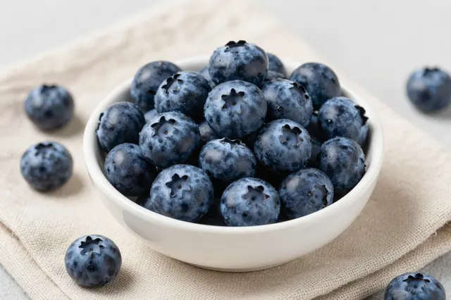 A small white bowl filled with ripe blueberries rests on a folded linen napkin, with extra berries scattered nearby.