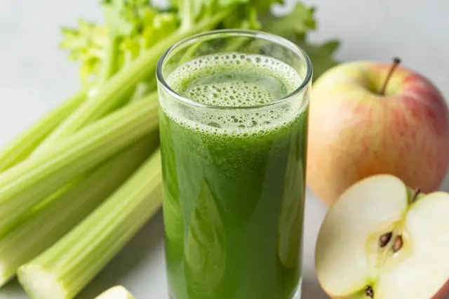 A glass of frothy green juice sits next to whole celery stalks and fresh apples on a light countertop