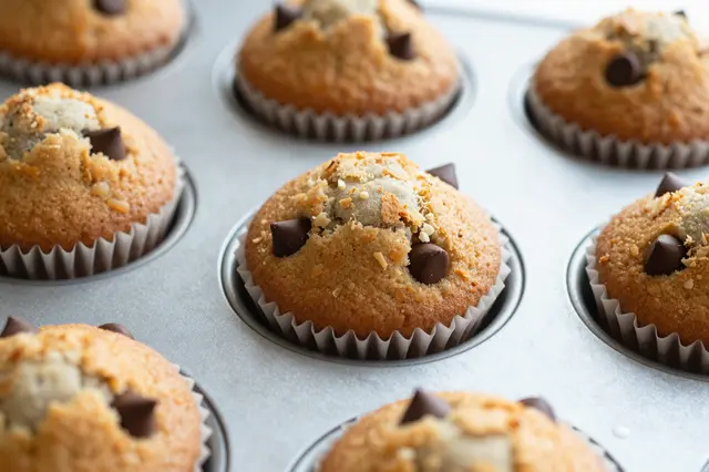 Warm domed chocolate chip muffins in paper liners sit in a metal baking tin