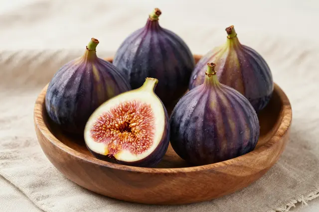 Whole and halved purple figs sit in a wooden bowl atop textured linen fabric.