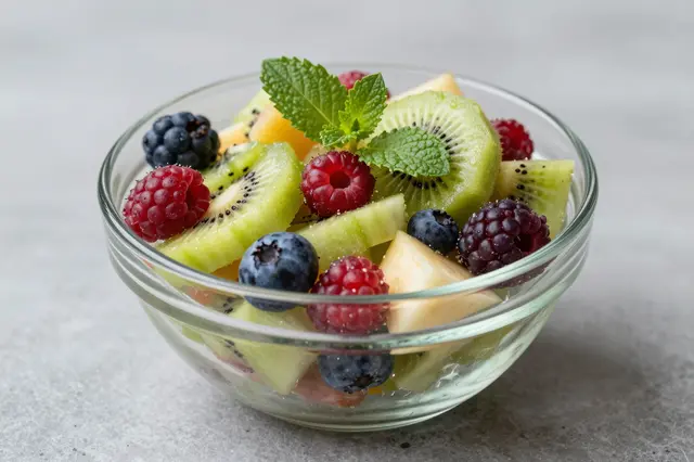 A clear glass bowl holds a vibrant fruit salad of kiwi, berries and fresh mint on a light gray countertop