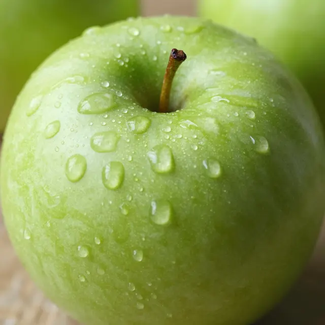 Close-up of a crisp green apple covered in clear water droplets, with blurred green apples in the soft background.