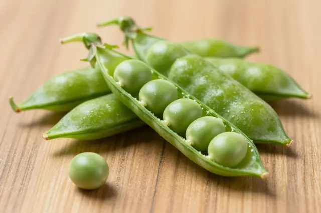 Open and closed fresh pea pods with one loose pea resting on a light wooden tabletop