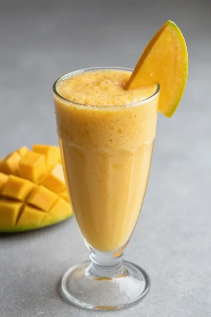 Tall glass of creamy mango smoothie garnished with a mango wedge, with cubed mango in the background on a gray surface
