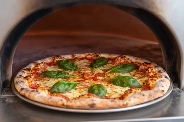 Freshly baked basil-topped pizza sits on a metal tray at the opening of a wood-fired oven.