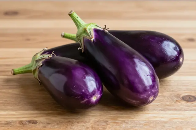 Three glossy fresh purple eggplants resting on a light wooden table surface