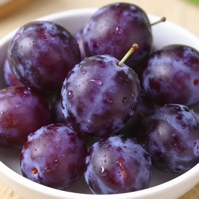 Ripe purple plums dotted with water droplets sit clustered in a white bowl