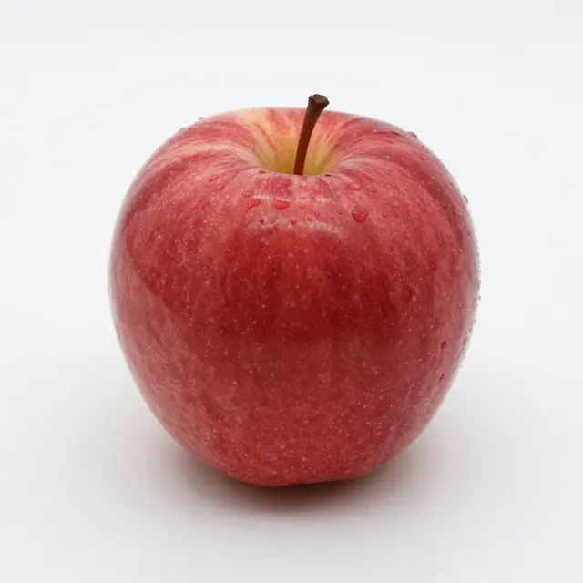 A whole ripe red apple with water droplets and a brown stem sits on a plain white background