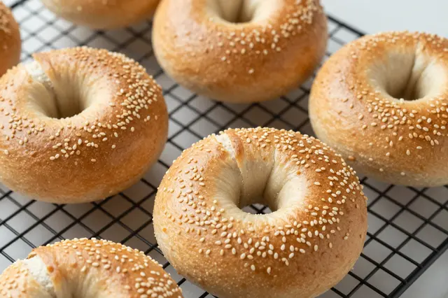 Golden sesame seed bagels rest on a black wire cooling rack over a light countertop