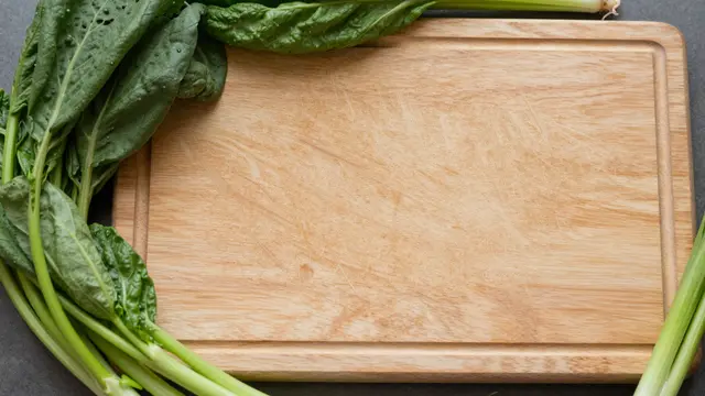 Top-down view of an empty wooden cutting board framed by leafy green vegetables on a dark countertop.
