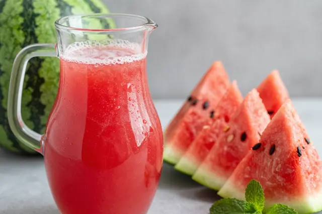 Glass pitcher of chilled watermelon juice sits next to sliced watermelon and fresh mint on a gray countertop