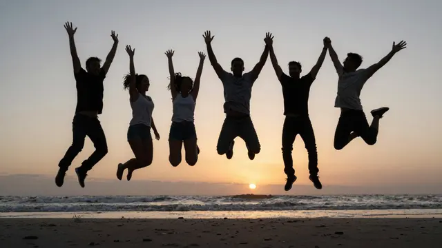 Silhouetted group of adult friends leap together above a sandy beach at golden sunset over the ocean