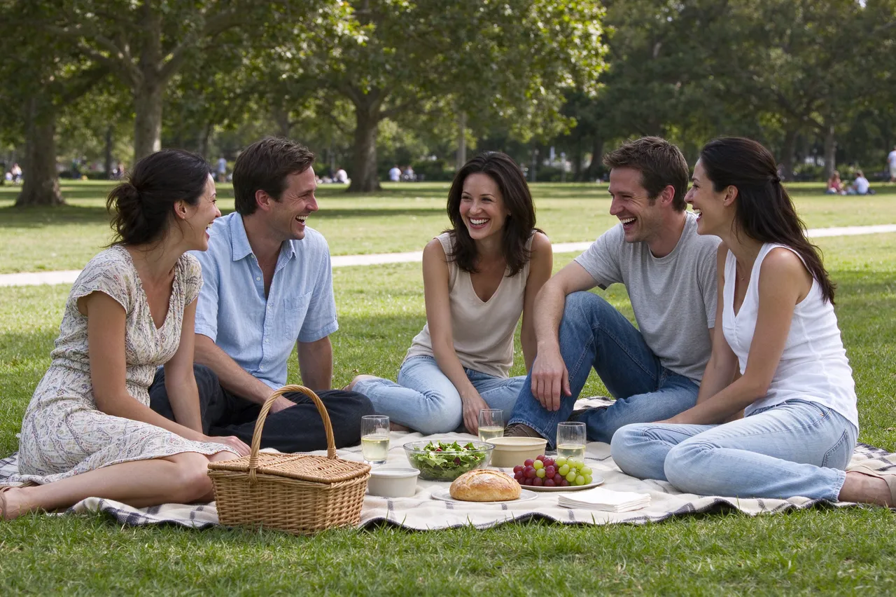 Five adult friends laugh together while sitting on a picnic blanket in a sunlit park