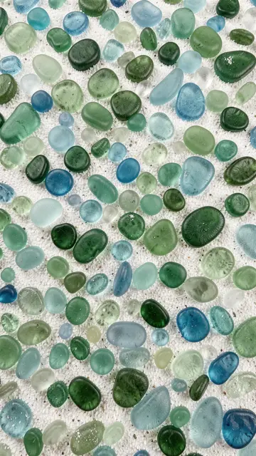 Smooth frosted green and blue sea glass pebbles scattered across fine white sand.