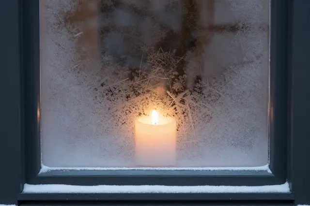 A lit candle glows through intricate frost patterns on a snow-dusted cabin window