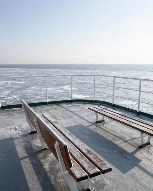 Empty frost-covered wooden benches sit on a ferry deck overlooking icy gray open water on a bright cold winter day