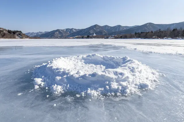A frozen lake with a central snow mound stretches toward distant snow-dusted mountains under a clear blue sky at midday.