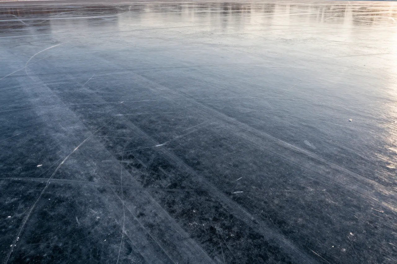 Clear dark ice of a frozen lake marked with long skate tracks under soft dawn light