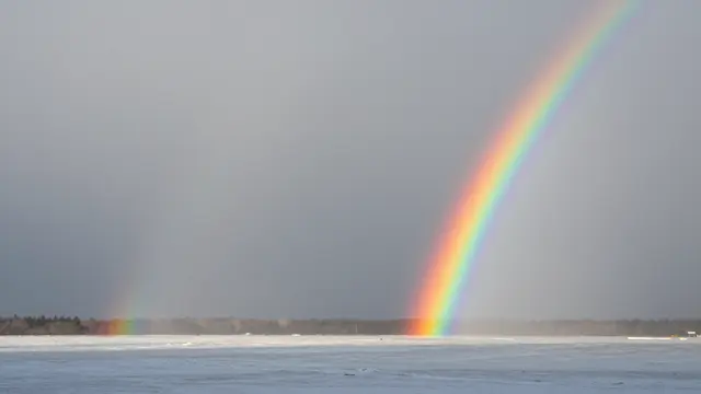 Two rainbow arcs stretch across a hazy gray sky over a frozen winter lake and distant treeline