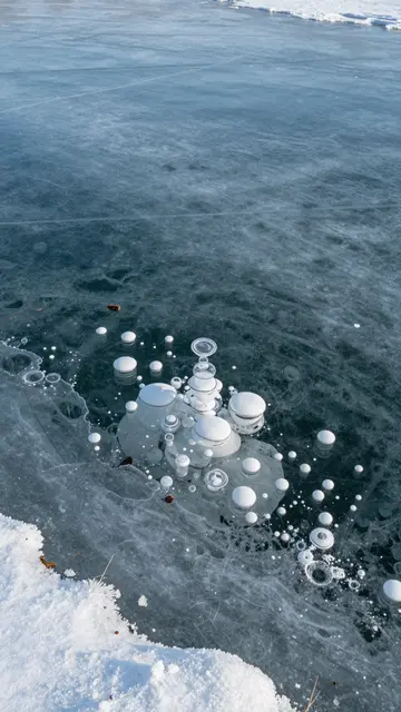 Translucent blue lake ice dotted with stacked trapped gas bubbles borders a snowy shoreline.