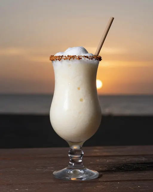 Frozen pina colada cocktail with a spiced rim rests on a wooden table against a beach sunset.