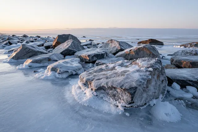 Ice-covered boulders sit along a frozen seashore lit by soft pale sunrise light