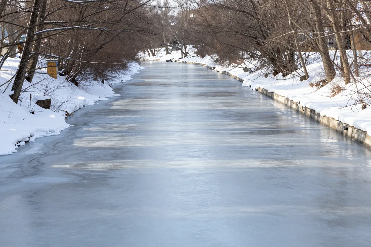 A partially frozen canal stretches between snow-covered banks lined with bare leafless winter trees.