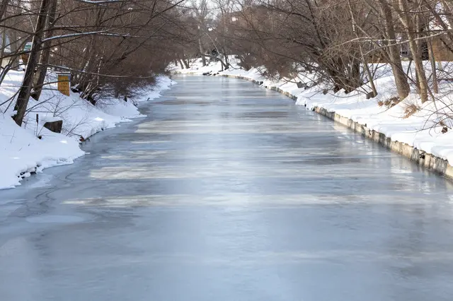 A partially frozen canal stretches between snow-covered banks lined with bare leafless winter trees.