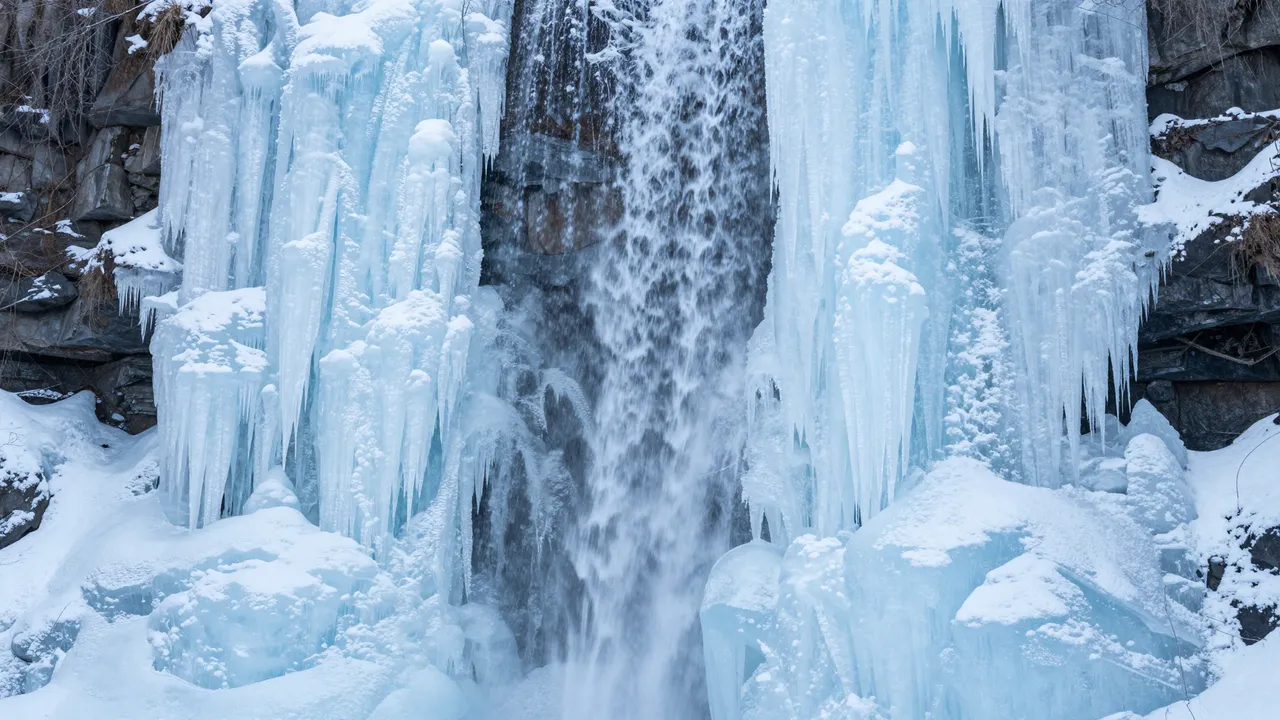 Partially frozen waterfall framed by pale blue icicles and snow-dusted dark rock in winter