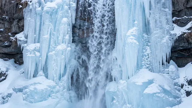 Partially frozen waterfall framed by pale blue icicles and snow-dusted dark rock in winter