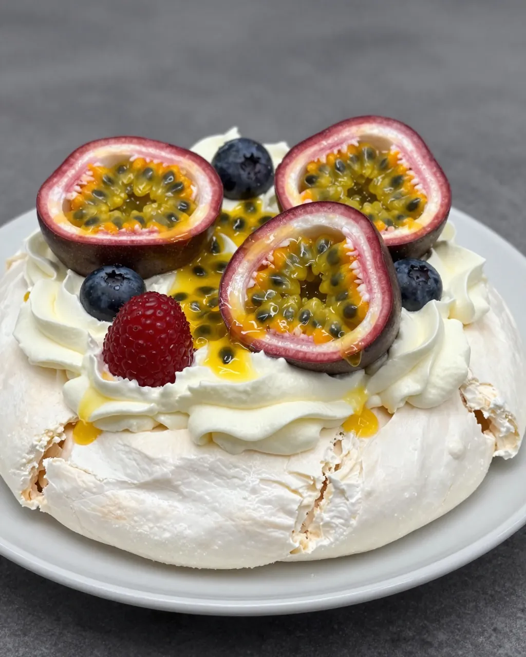 A meringue pavlova topped with whipped cream, passion fruit halves, blueberries and a raspberry rests on a light plate.