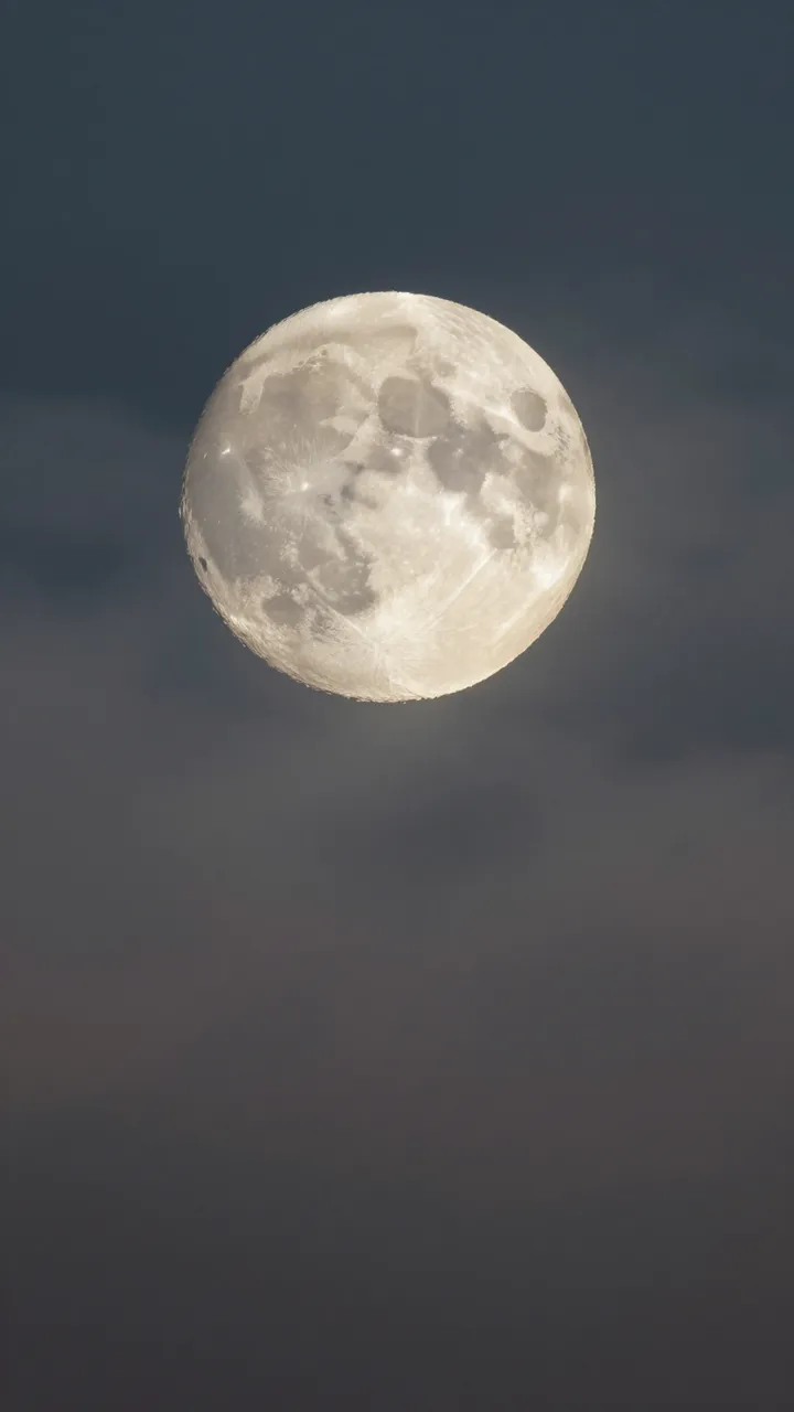 A detailed full moon sits against a hazy, dark twilight night sky with soft drifting clouds.