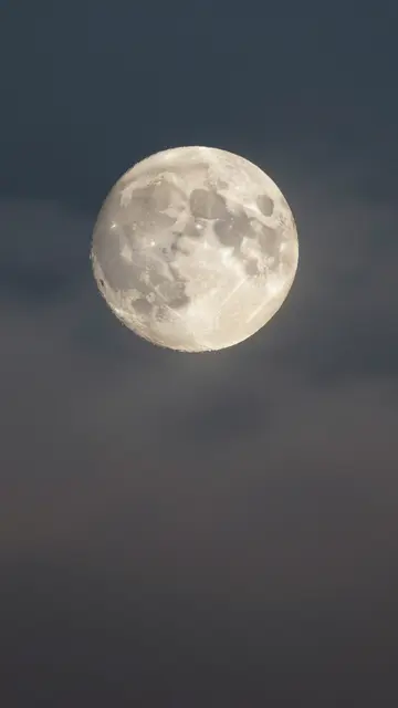 A detailed full moon sits against a hazy, dark twilight night sky with soft drifting clouds.