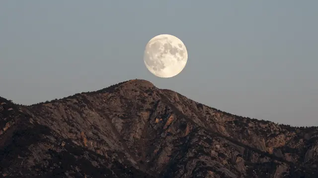 A large full moon hovers just above a rugged rocky mountain peak at twilight