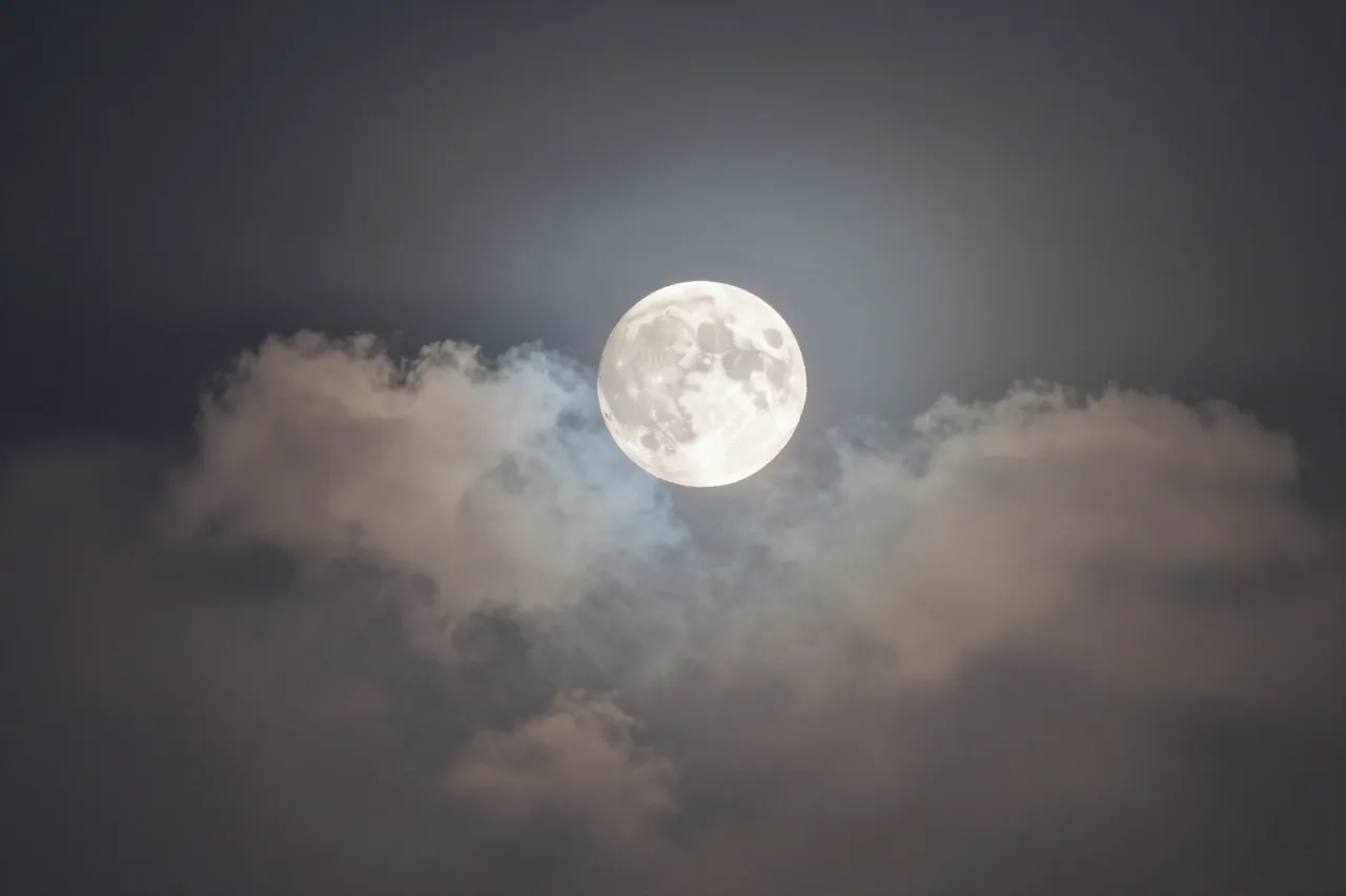 Bright full moon glows with a soft halo above wispy moonlit clouds in a dark night sky