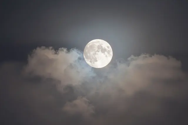 Bright full moon glows with a soft halo above wispy moonlit clouds in a dark night sky