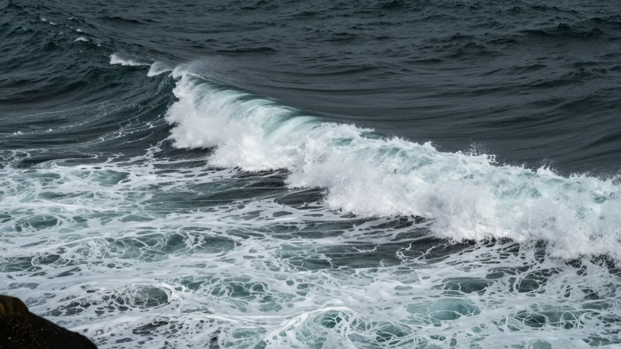 A cresting foam-streaked ocean wave rolls through dark choppy gale sea viewed from a cliff edge