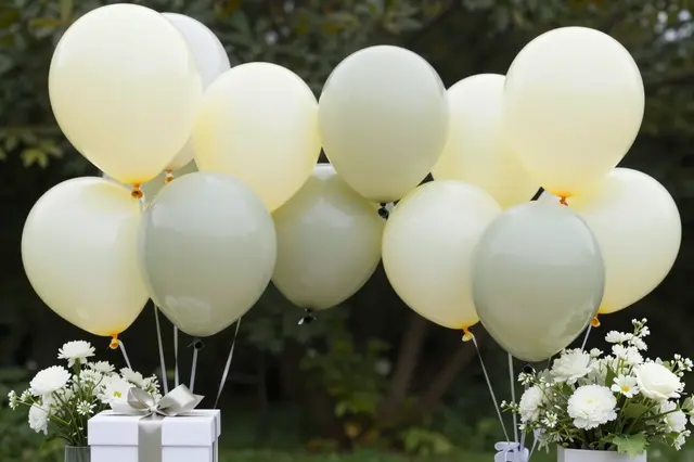 Pale yellow and sage balloons flank a gift box and white flower arrangements in an outdoor garden