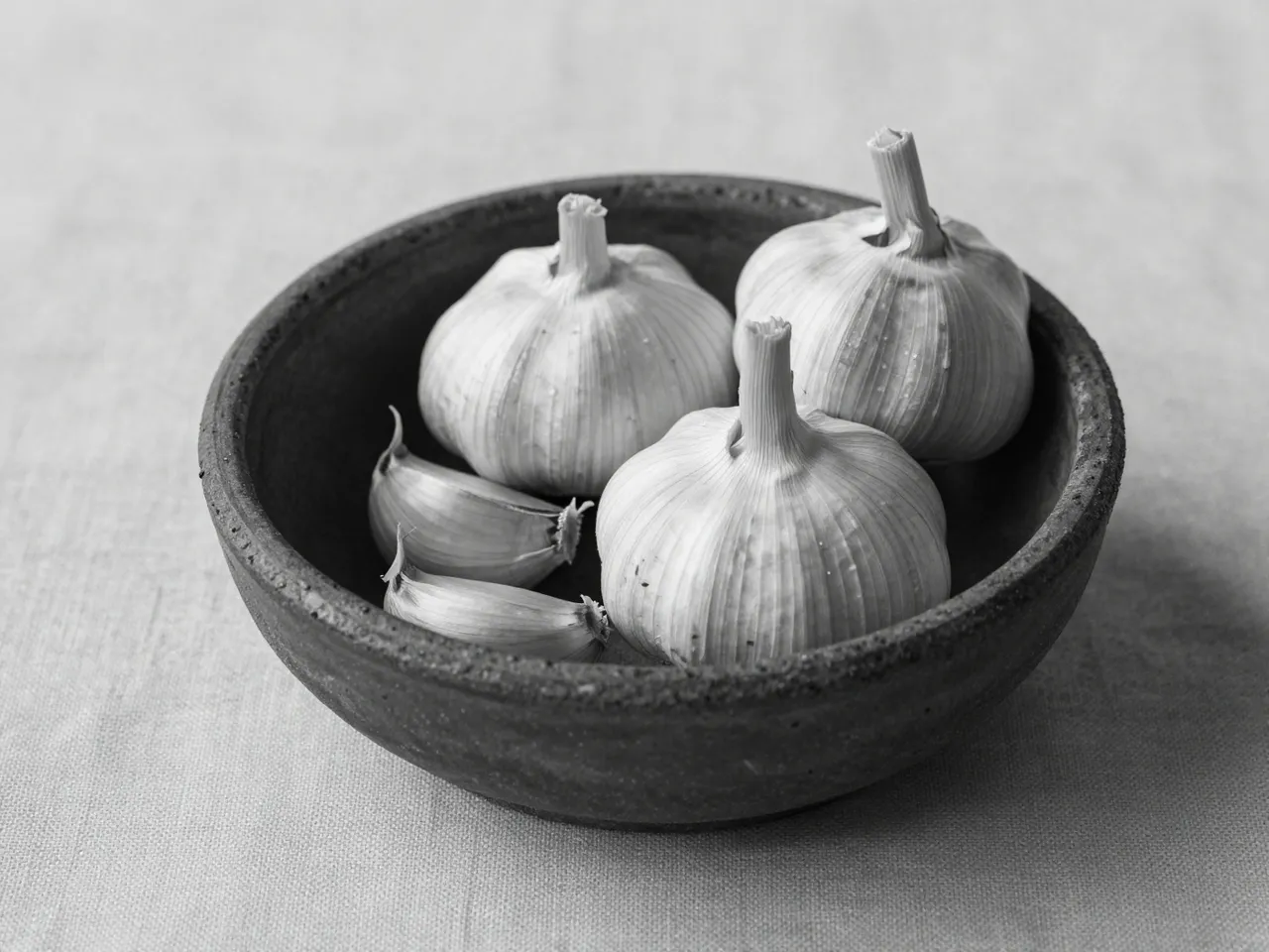 Black and white still life of whole garlic bulbs and cloves in a rough clay bowl on linen fabric