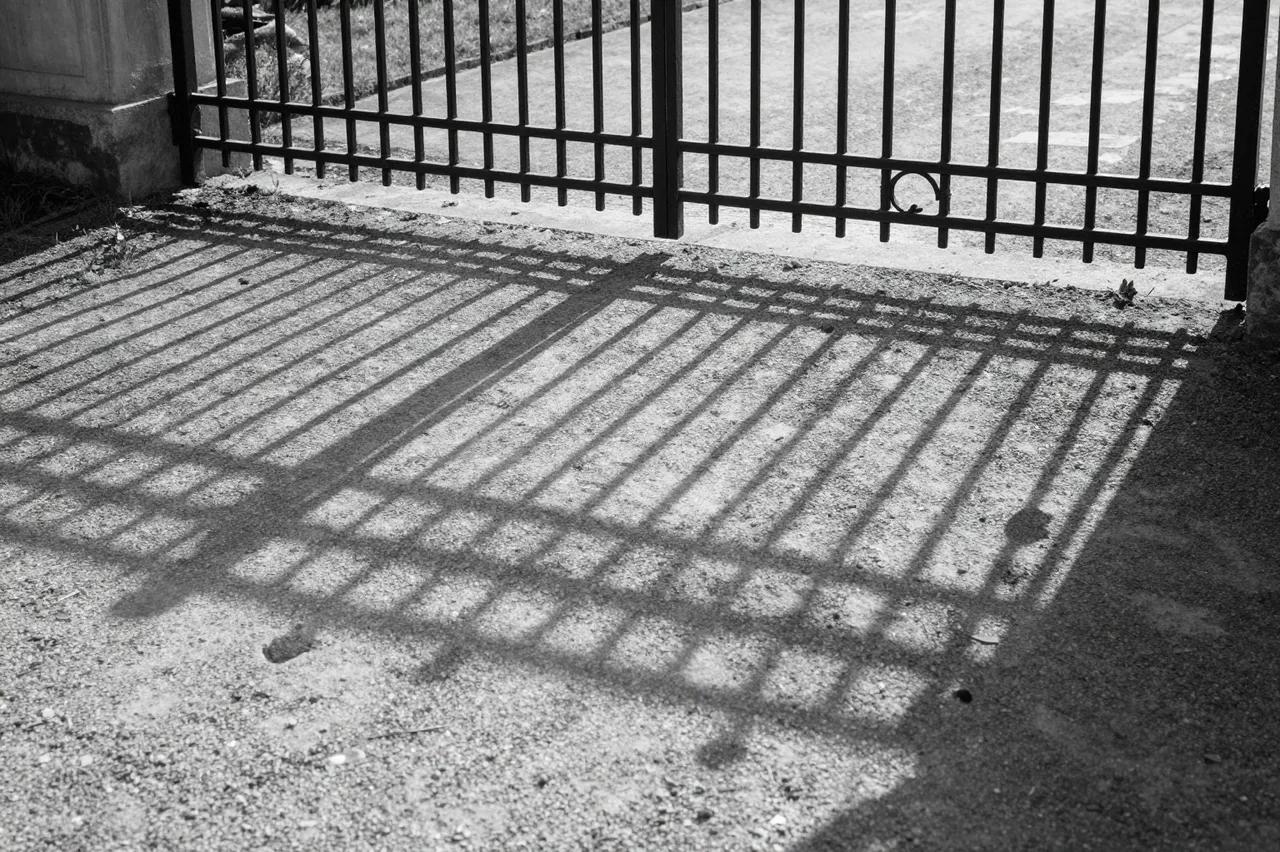 Black and white shot of a wrought iron gate's geometric shadow stretching across raked gravel in low light