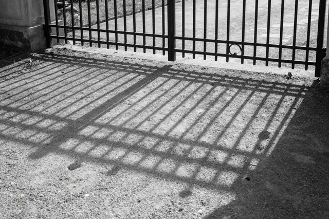 Black and white shot of a wrought iron gate's geometric shadow stretching across raked gravel in low light