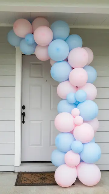 A pale pink and powder blue balloon arch frames a white front porch door for a gender reveal celebration.