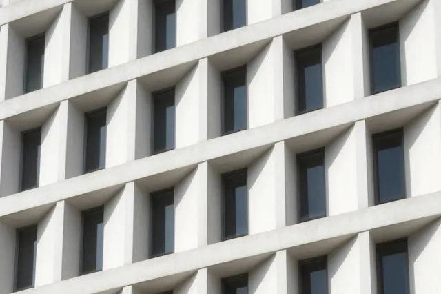 Close-up of a modern white geometric building facade with sharp sunlit shadows around recessed windows.
