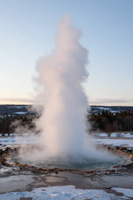 A geyser erupts with a tall steam column in a snow-dusted valley at soft dawn light