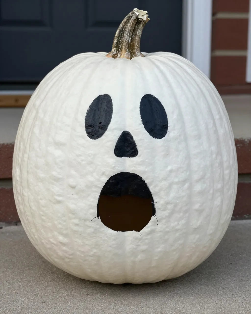 A white pumpkin painted as a spooky friendly ghost sits on a concrete front porch step