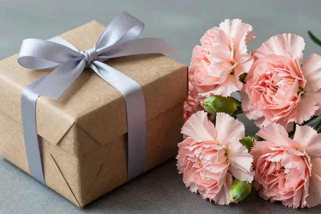 Brown kraft gift box tied with gray satin ribbon sits next to a bouquet of soft pink carnations on a smooth gray surface.