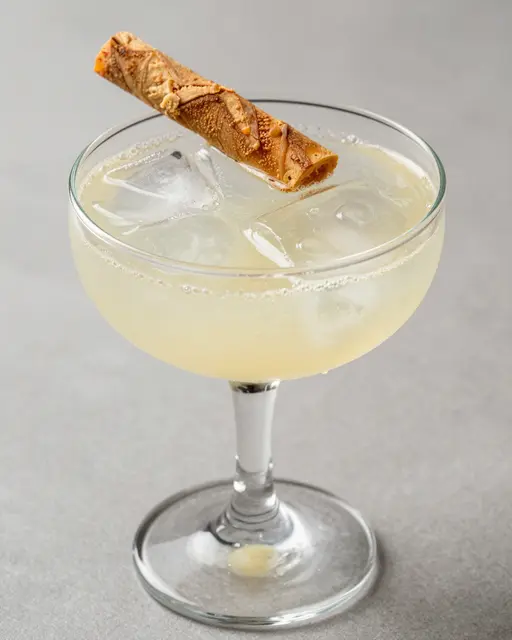 A pale bubbly ginger lemon fizz mocktail with ice and a rolled garnish in a coupe glass on a neutral gray surface
